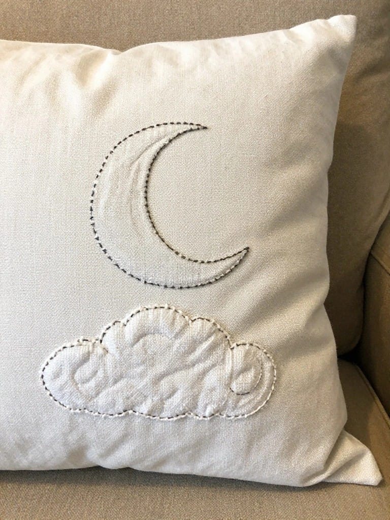 White pillow cover embroidered with a gray-outlined crescent moon and a puffy white cloud below it on a beige couch.