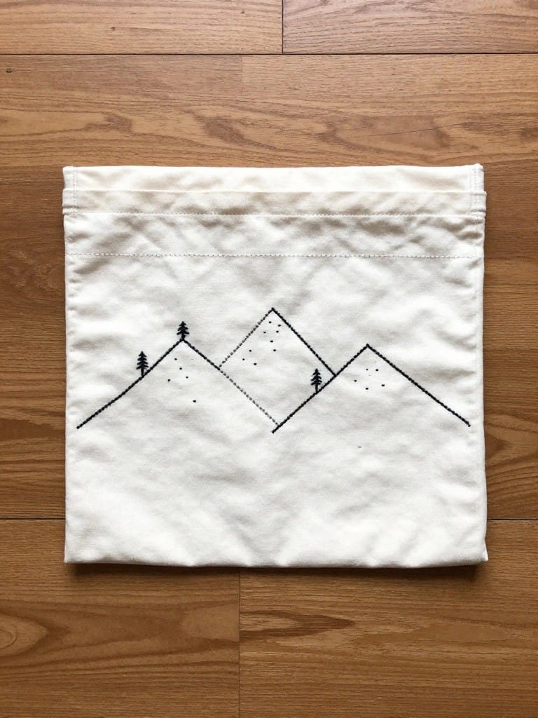 White drawstring pouch with black embroidered line art of three mountains, pine trees, and dots on the front.