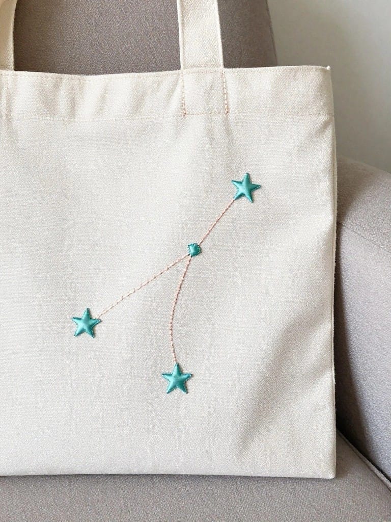White canvas tote bag embroidered with turquoise stars and lines forming the Cancer constellation on one side.