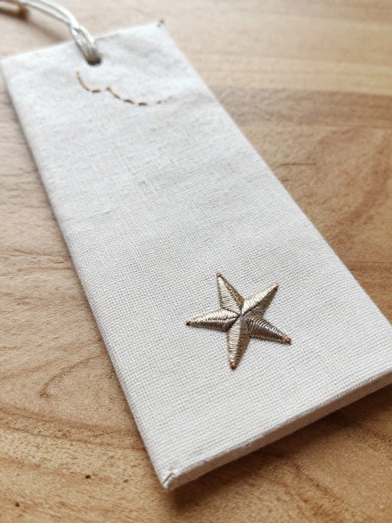 White linen bookmark with gold embroidered star near bottom edge and small curved moon above it, tassel strings at top.