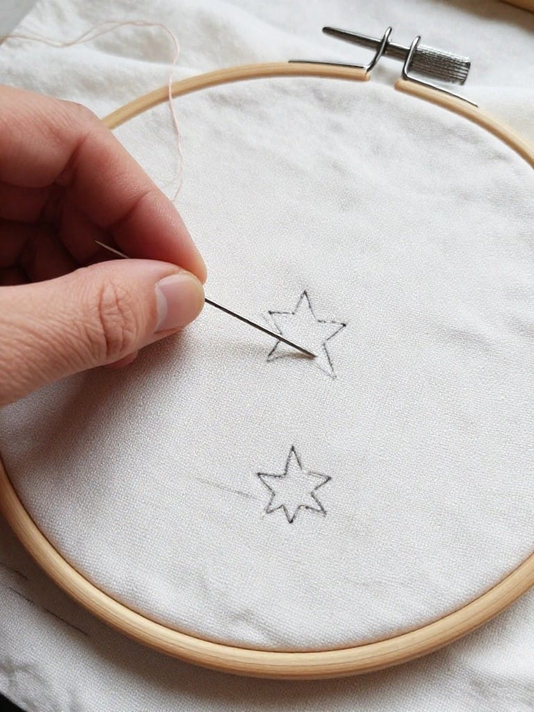 Hand holding a needle stitching a black outline on a partial five-point star, with another outlined star below it, on white fabric in a wooden embroidery hoop.
