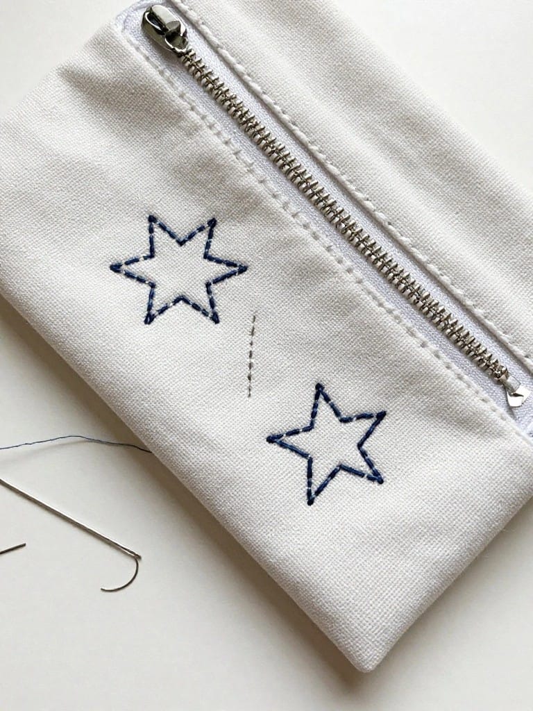 White zippered canvas pouch embroidered with navy blue outline stars in different sizes, needle and thread nearby.