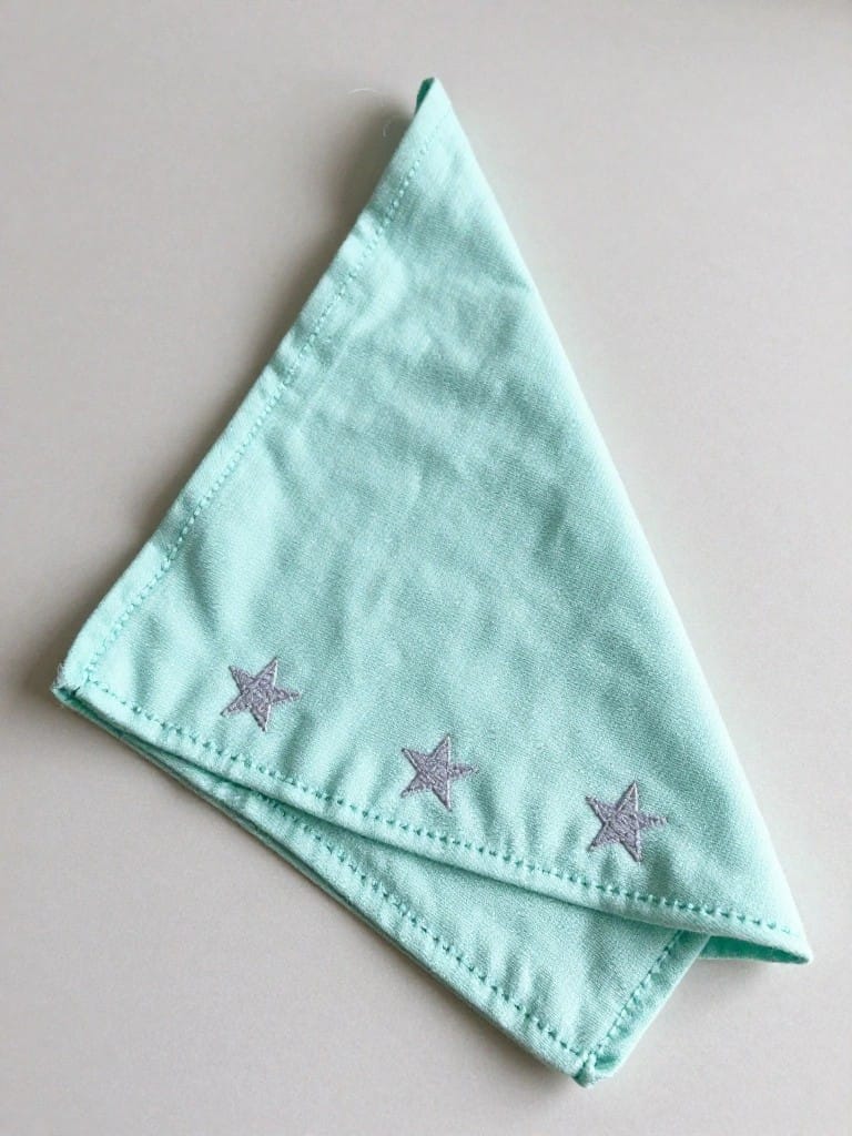 A mint green cloth napkin folded into a triangle with three small silver embroidered stars in a row along one slanted edge.