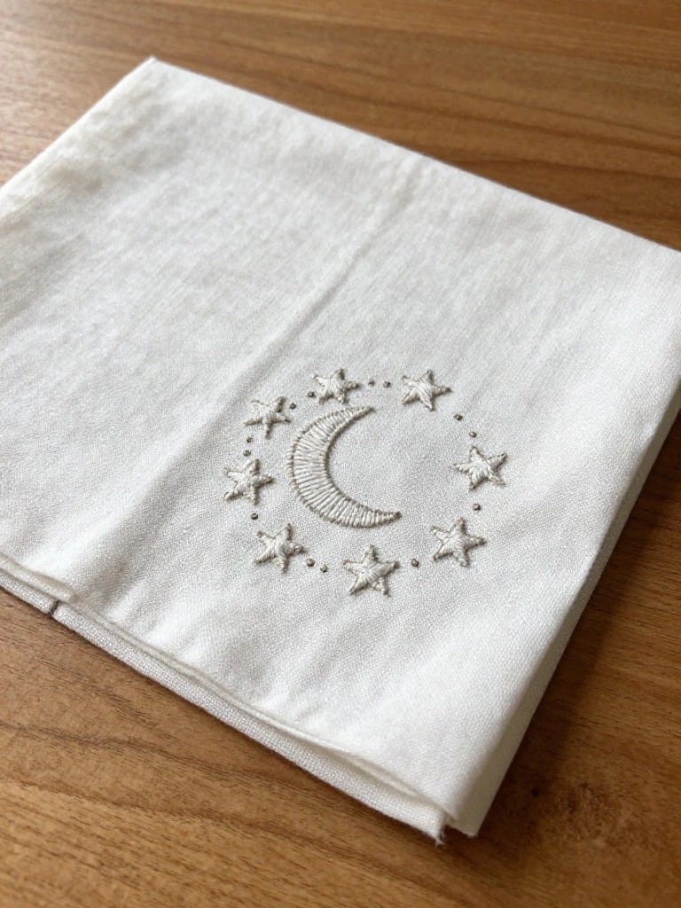 White napkin embroidered with light gray crescent moon centered in a circle of stars.