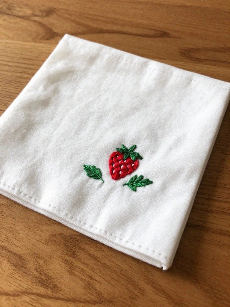 White square napkin with small embroidered red strawberry and green leaves in one corner on wooden table.