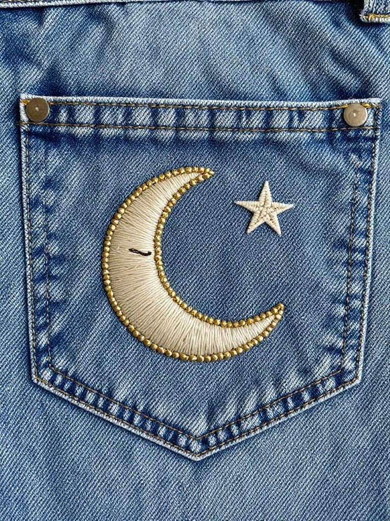 Denim jeans back pocket embroidered with a white crescent moon outlined in gold thread and a small white star next to it.