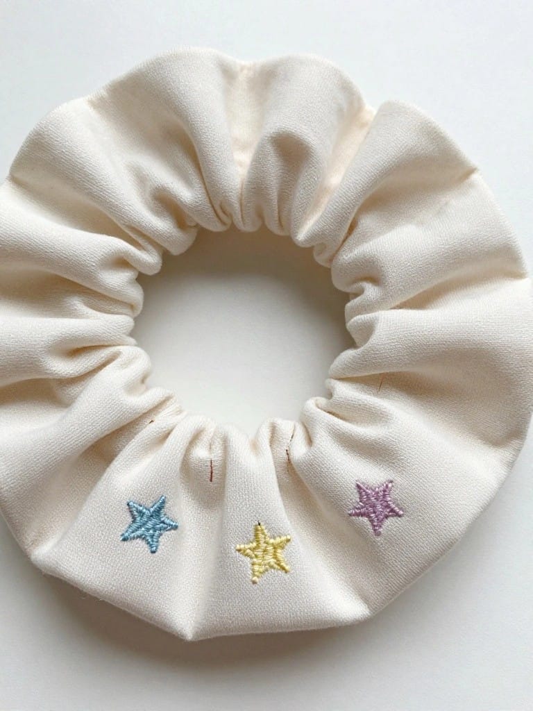 White fabric scrunchie embroidered with three small stars in blue, yellow, and purple spaced around the band.