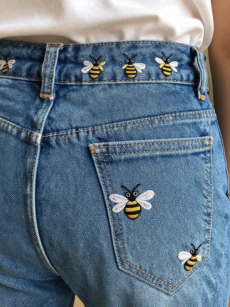 Blue denim jeans feature black and yellow embroidered bees in a row along the waistband and clustered on the back pocket.