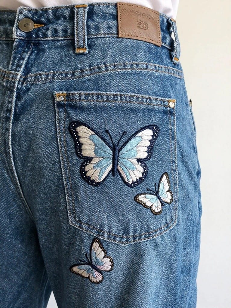 Back pocket of blue jeans embroidered with one large blue and white butterfly in the center and three smaller ones around it on the denim.