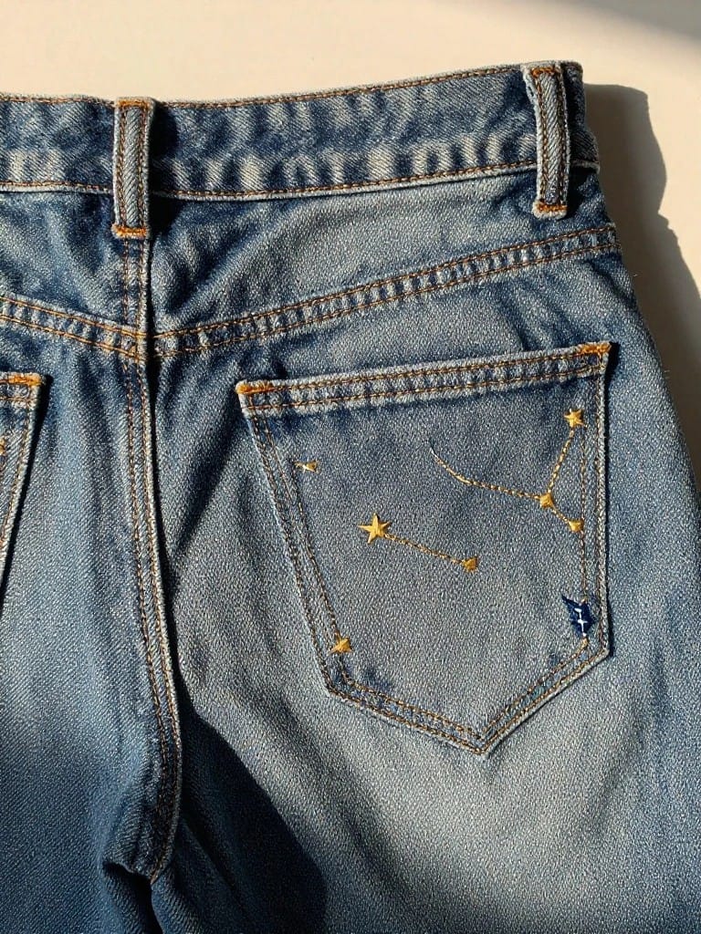 Back pocket of light wash jeans with yellow embroidered stars connected into a constellation and a small blue butterfly below.