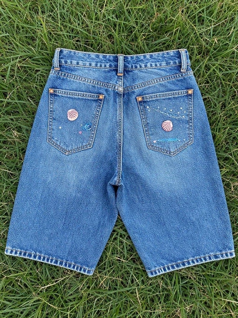 Back view of cropped blue denim jeans with embroidered pink seashells and sparkles on the pockets, lying flat on grass.