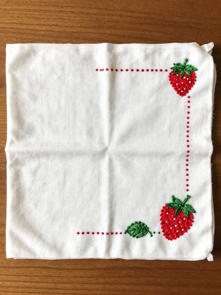 White square napkin with red dashed border embroidery and small strawberry motifs with green leaves in the corners.