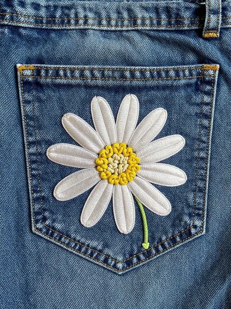 White daisy with yellow center and green stem embroidered on the back pocket of blue jeans.