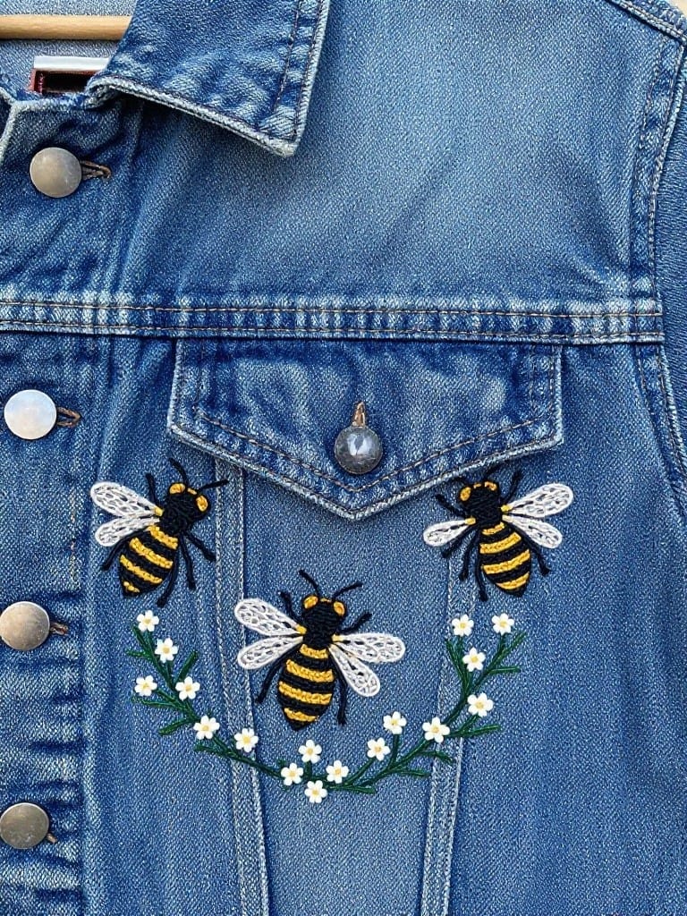 Denim jacket pocket embroidered with three yellow and black bumblebees in a circular arrangement surrounded by small white flowers and green leaves.
