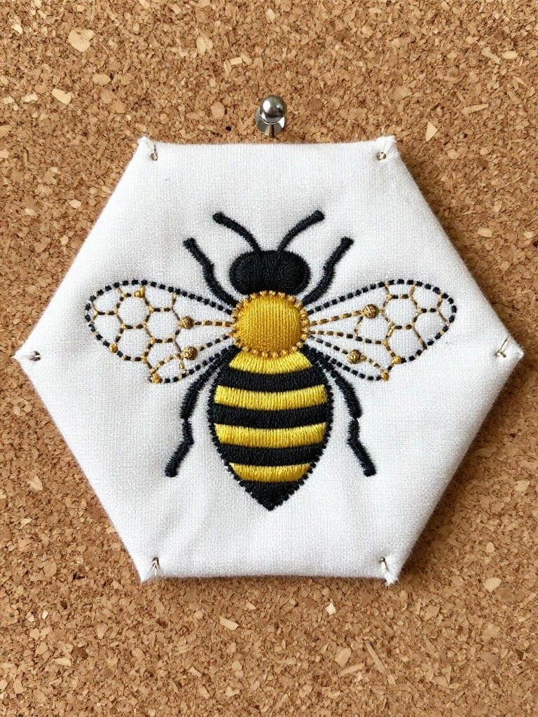 White hexagonal fabric with black-and-yellow embroidered bee and gold accents pinned to a corkboard.