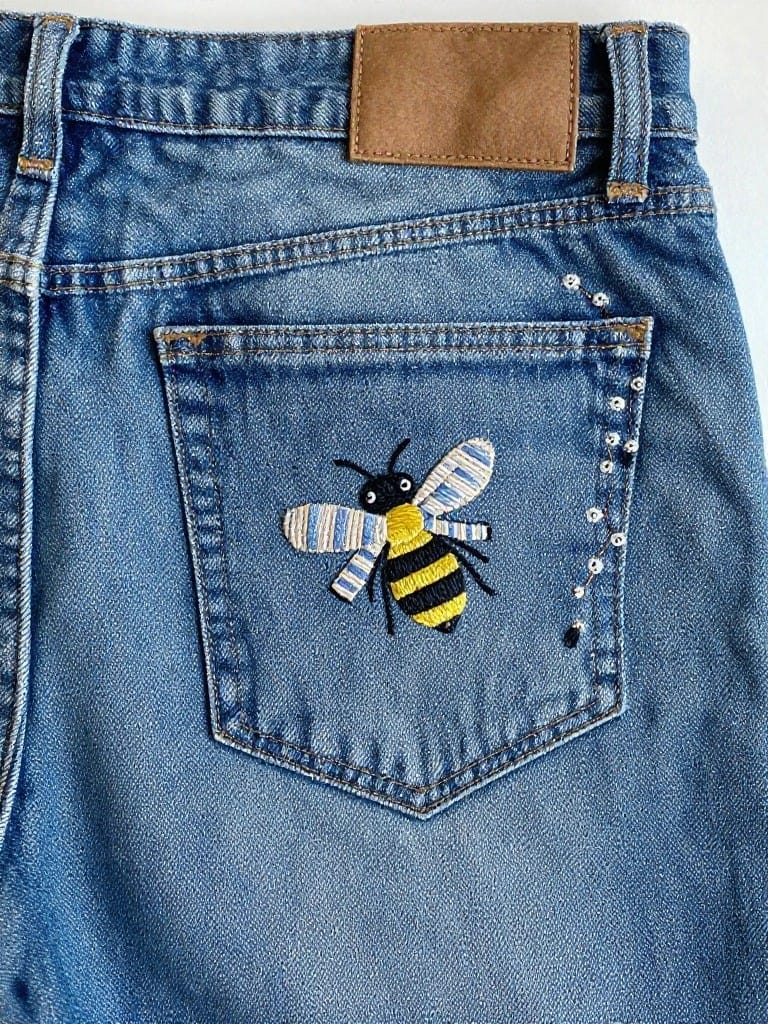 Black and yellow embroidered bee with striped wings and surrounding white dots on the back pocket of blue jeans.