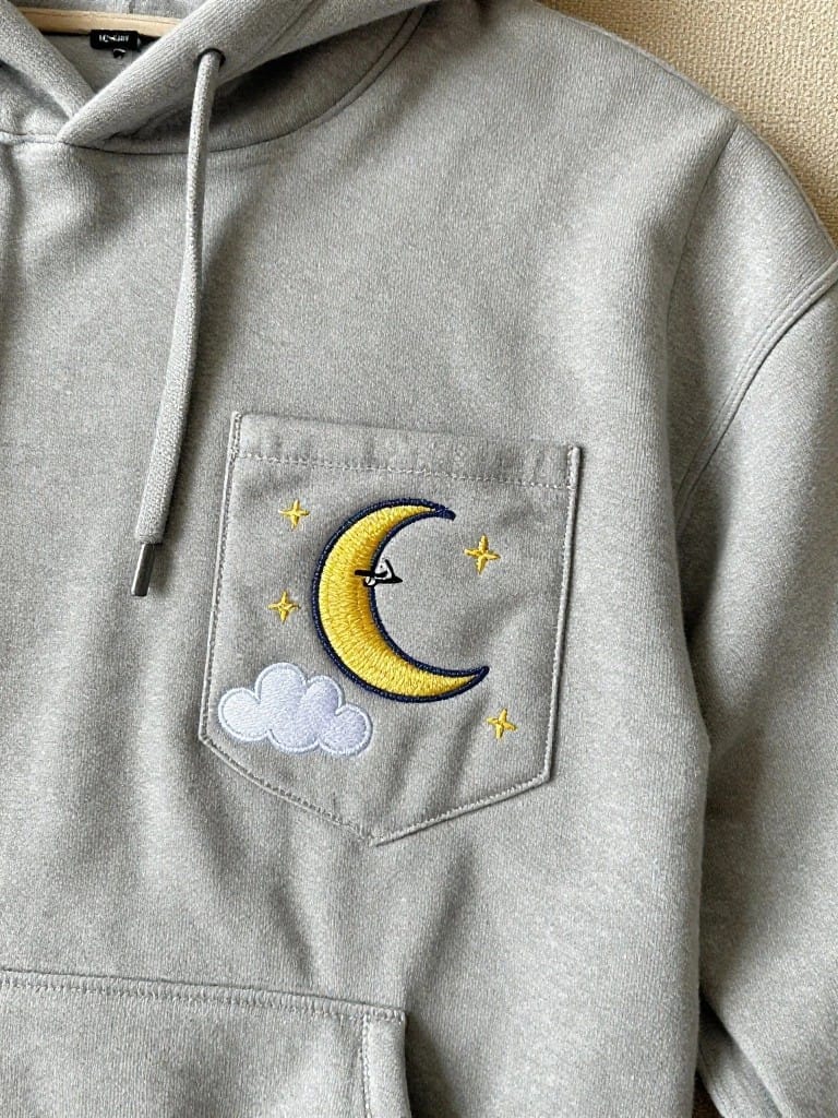 Gray hoodie with yellow embroidered crescent moon containing an airplane, plus stars and cloud on the chest pocket.