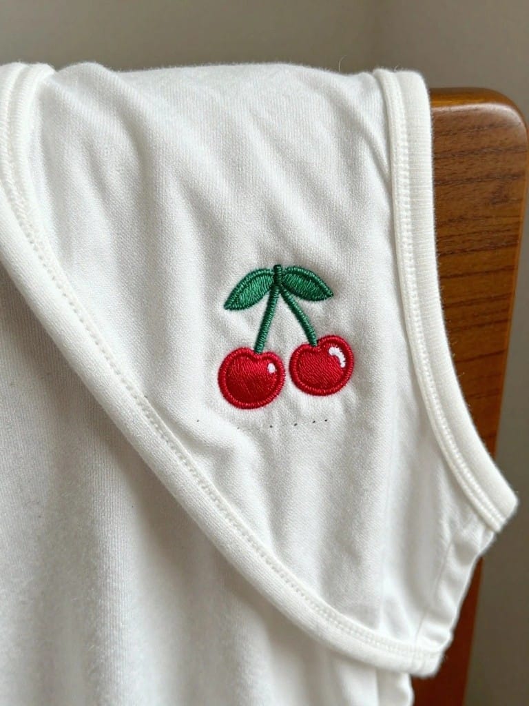 White sleeveless onesie with red embroidered cherries and green leaves on the upper left shoulder strap, draped over a wooden chair back.