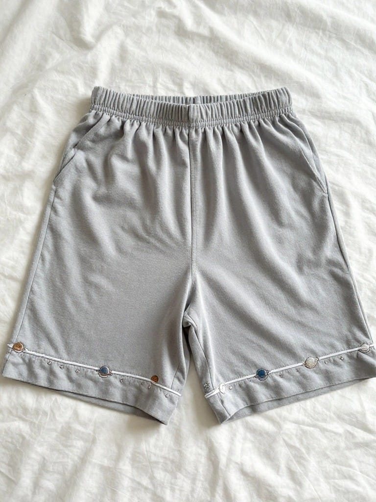 Light gray knit shorts with small orange and blue embroidered circles and white stitching along the bottom hems of both legs.