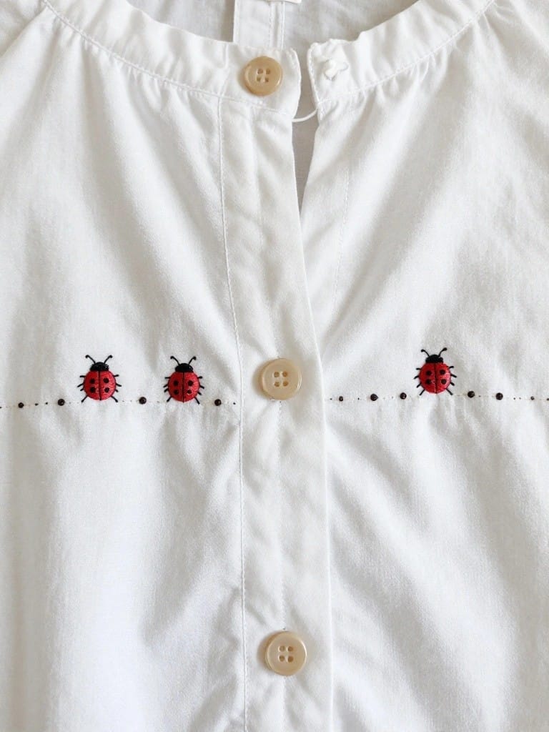 Close-up of embroidered red ladybugs with black spots and dotted trail along the button placket of a white child’s blouse.