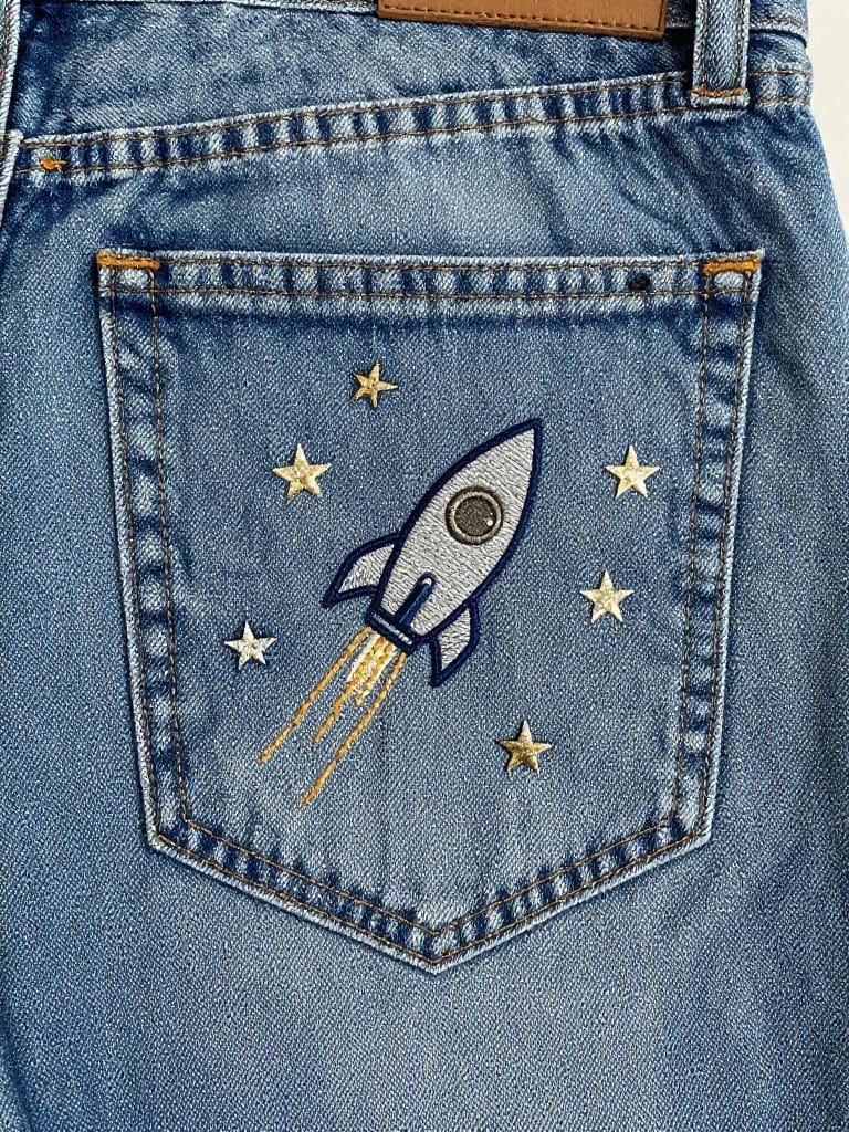 Back pocket of light wash jeans with embroidered white rocket ship, gold flame trail and stars.