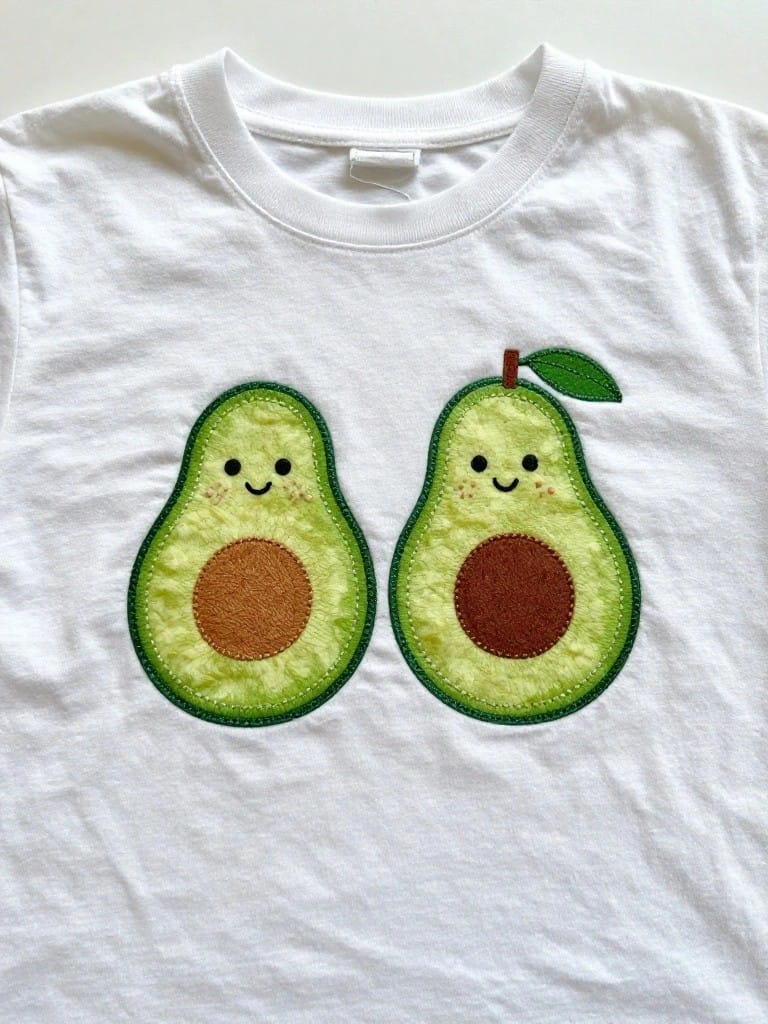White children's t-shirt embroidered with two green smiling avocados on the front chest area.
