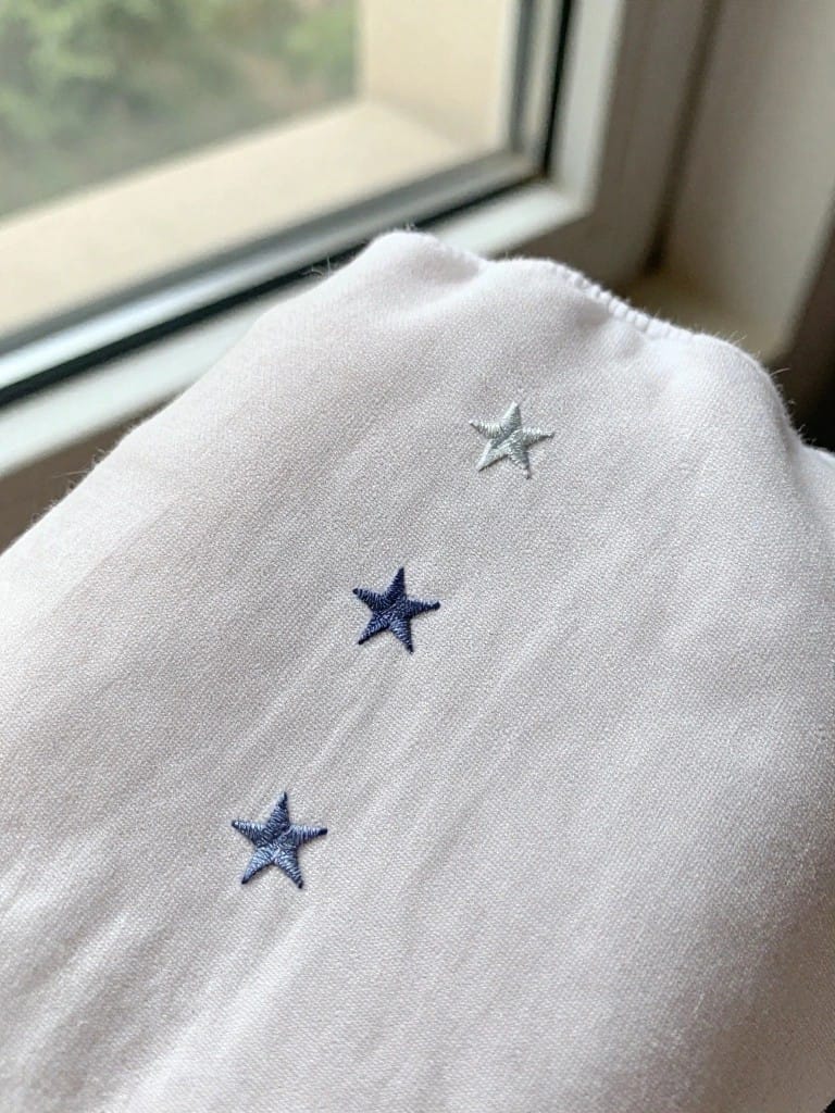 White blanket edge with three embroidered stars in light to dark blue shades aligned vertically.