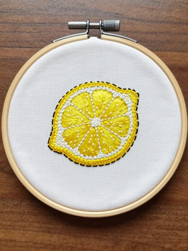 Small embroidery hoop on white fabric with a yellow lemon slice cross-section outlined in black.