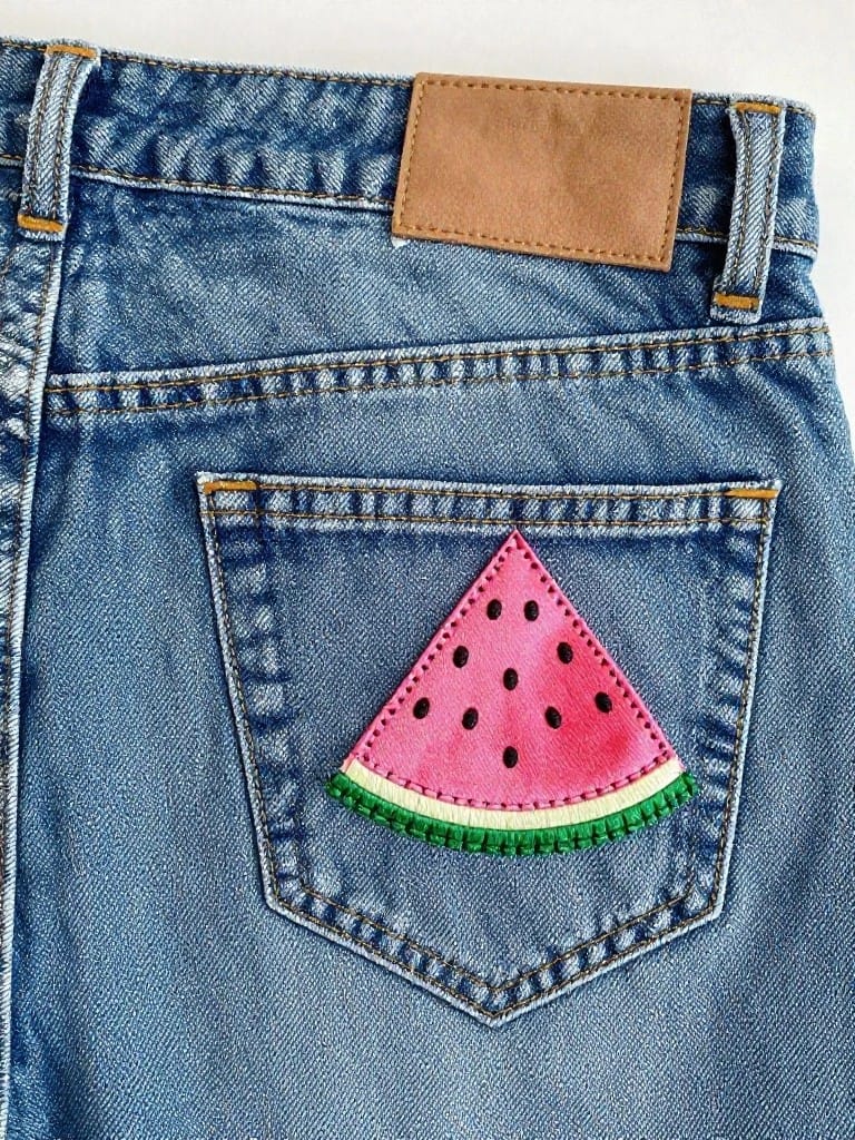 Back pocket of blue jeans with embroidered pink watermelon slice, black seeds, and green rind.