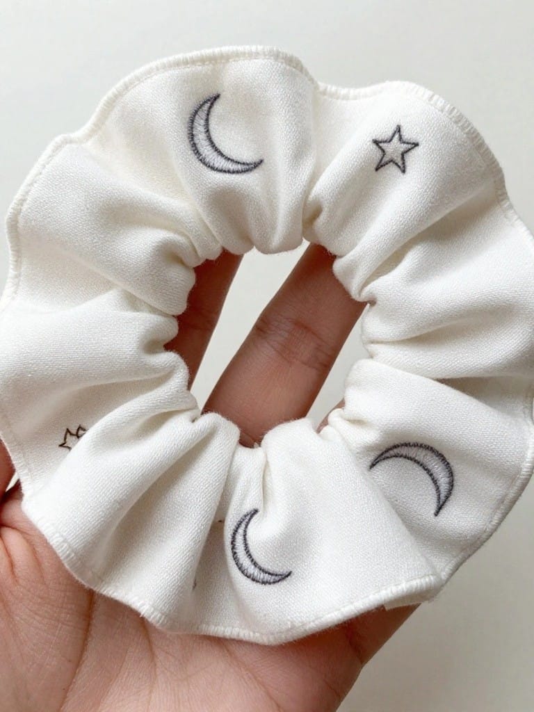 White scrunchie embroidered with gray crescent moons and stars, held in a hand.