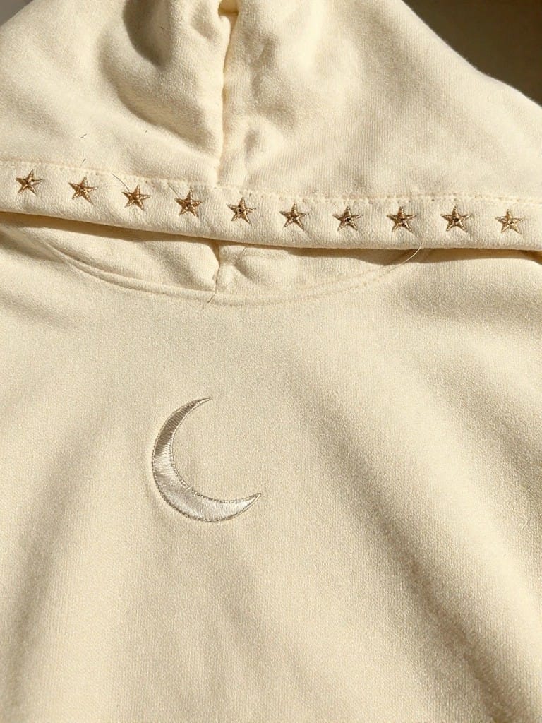 Cream hoodie features a white embroidered crescent moon on the chest and gold star embroidery along the hood edge.