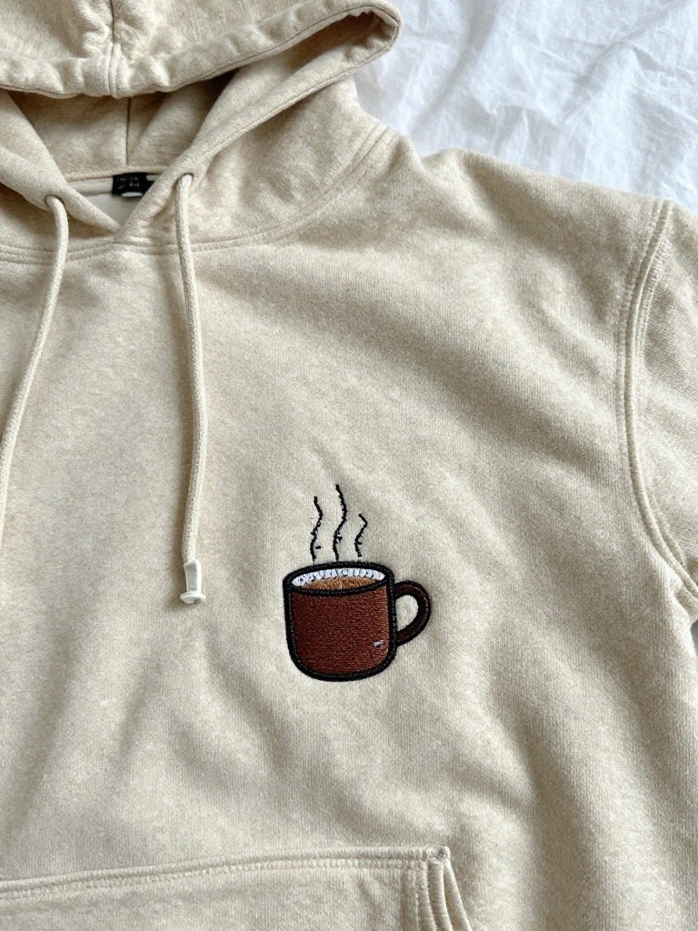 Beige hoodie with a small embroidered brown coffee mug and steam on the left chest.