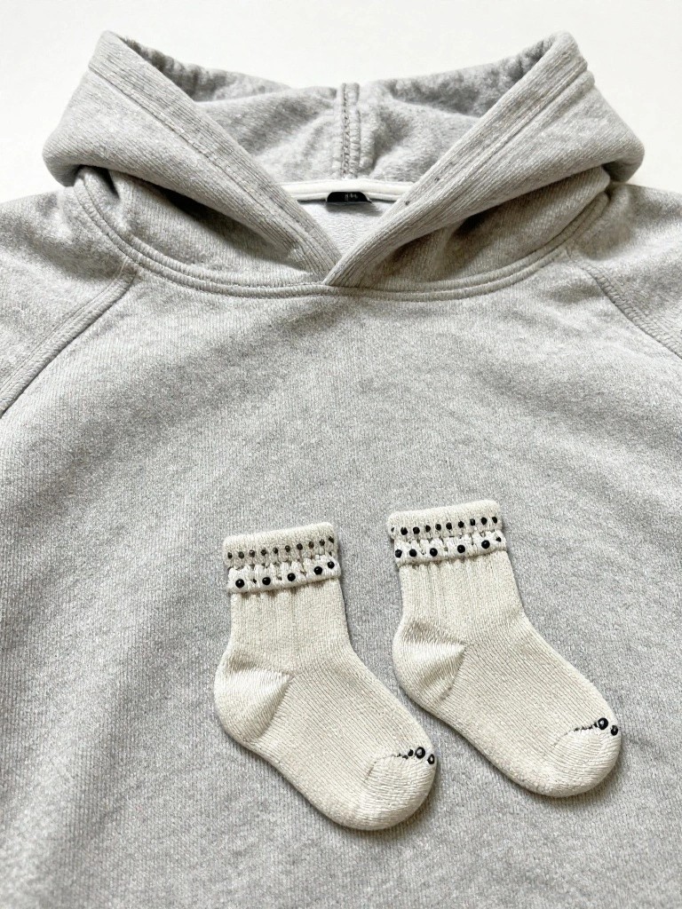 Gray hoodie laid flat with two small embroidered white baby socks, each with black-dotted cuffs, placed side by side on the front below the hood.