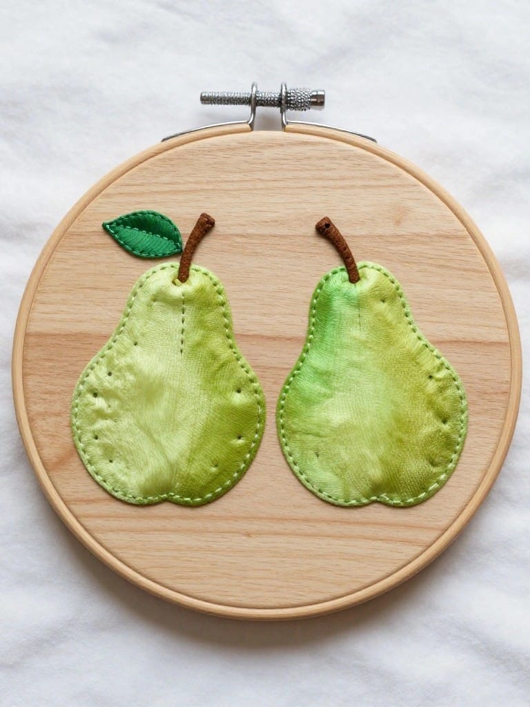 Wooden embroidery hoop with two green fabric pears, brown stems, and green leaves on white fabric.