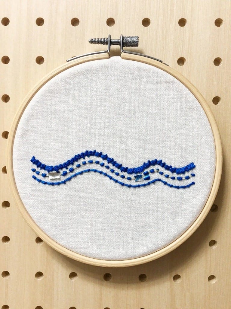 White embroidery hoop with four blue wavy lines stitched across the center, some with blue knots, mounted on wooden pegboard.