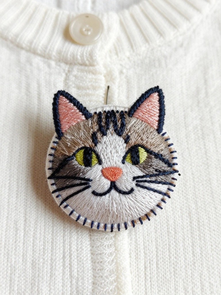 White knit cardigan with a circular embroidered tabby cat face brooch pinned near the collar.