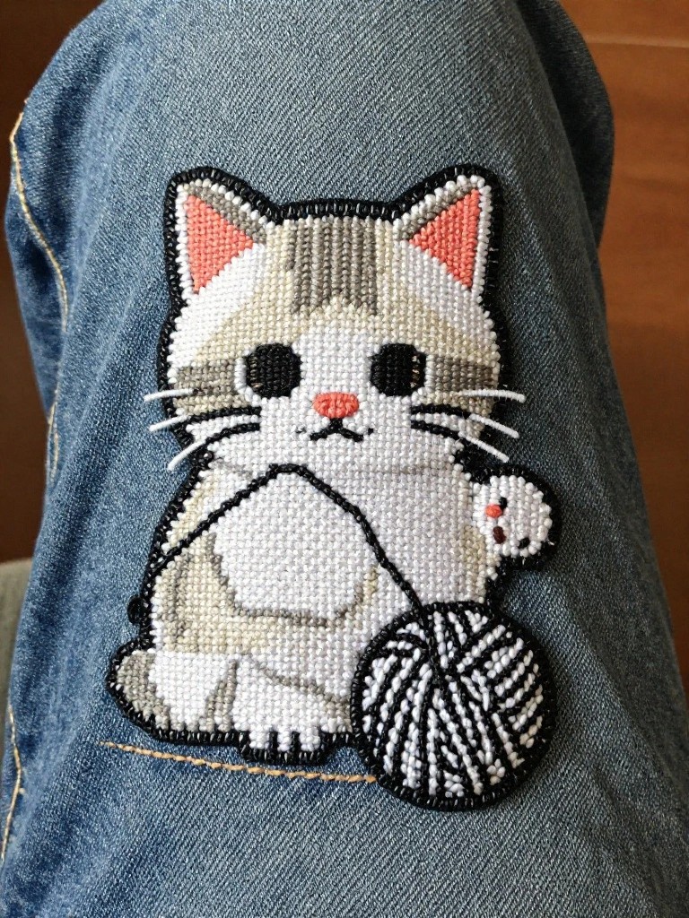 Embroidery patch of a grey and white kitten holding a yarn ball sewn onto the thigh of blue jeans.