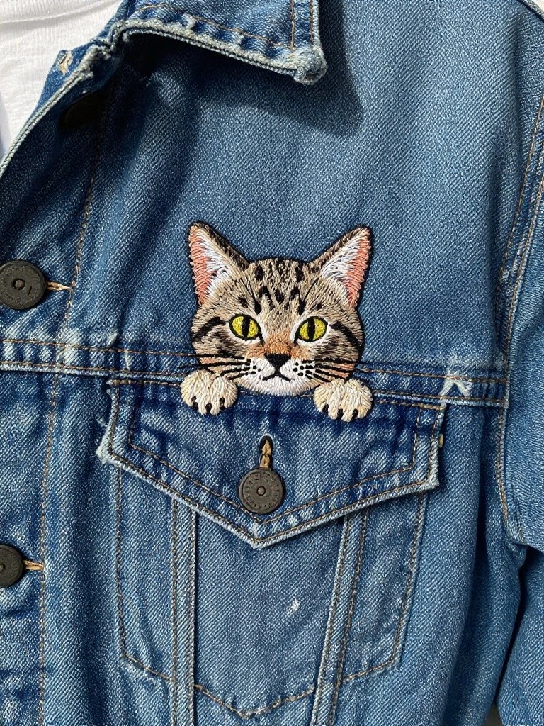 Denim jacket with embroidered tabby cat face and front paws on the edge of a chest pocket.