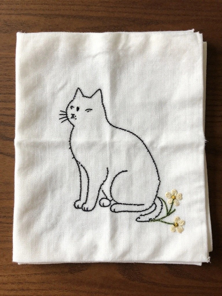 White folded napkins embroidered with a black outline cat sitting sideways and small yellow flowers emerging from behind it.