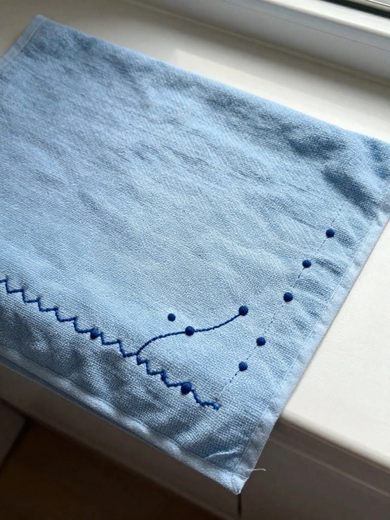 Blue terrycloth towel with blue embroidered wavy line and dots along one corner edge.