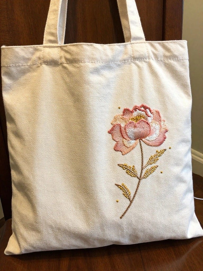 White canvas tote bag with pink peony flower, gold stem, and leaves embroidered on the lower front.