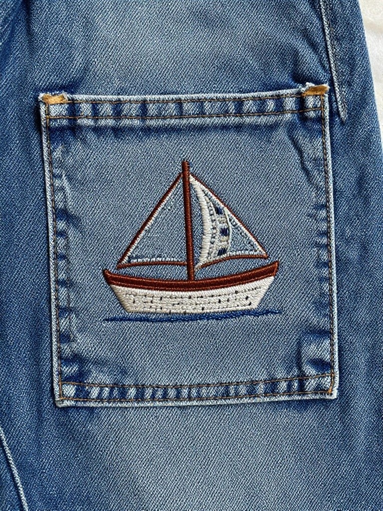 Small embroidered sailboat with sails and waves on the pocket of light blue denim jeans.