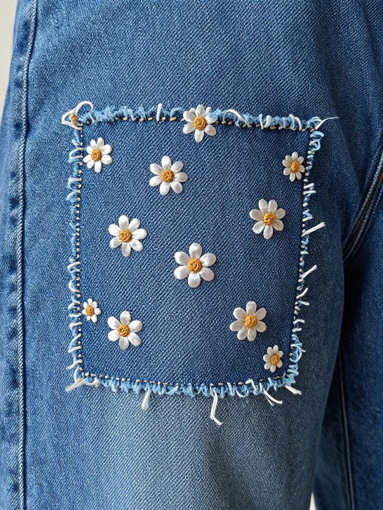 Blue jeans pocket with a square embroidered patch of small white daisies, yellow centers, and frayed edges.