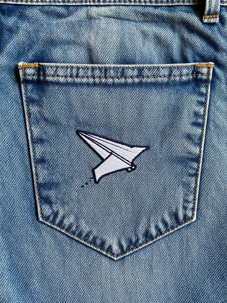 White embroidered paper airplane with dotted trail on the back pocket of blue jeans.