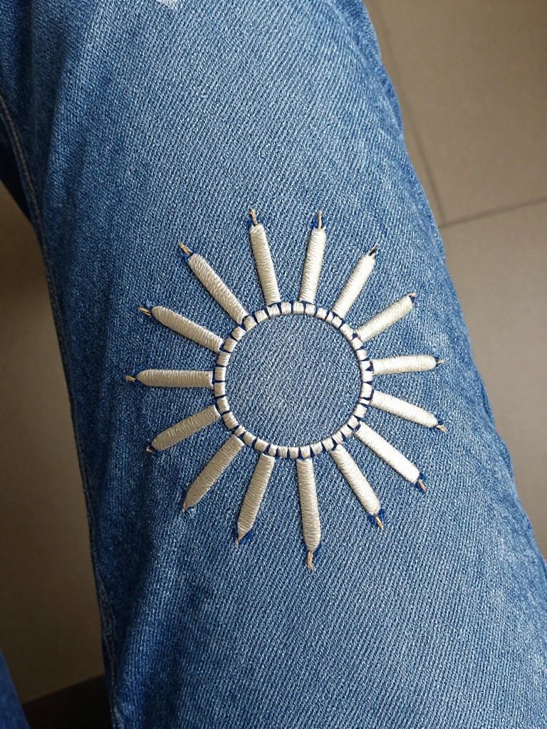 White embroidered sun with a central circle and sixteen tapered radiating rays on the blue denim leg of jeans.