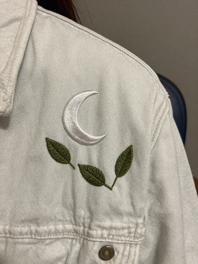 White crescent moon and three green leaves embroidered on the shoulder of a light denim jacket.
