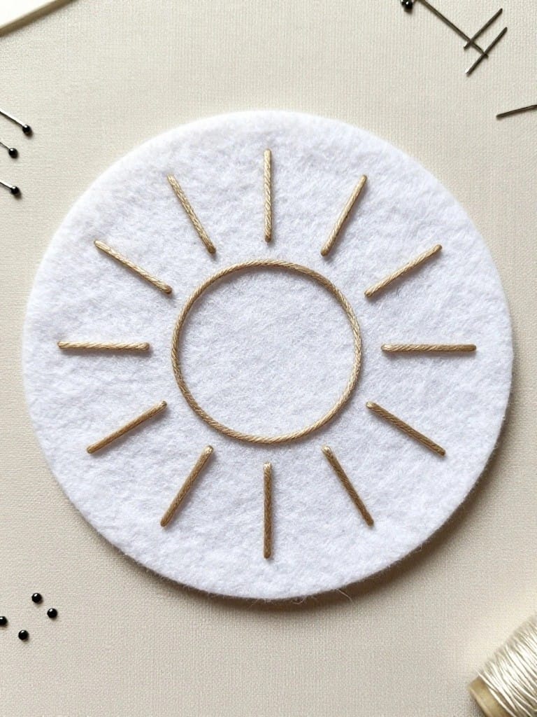 White felt disc embroidered with gold sun motif of central circle and radiating rays, pins securing stitches, gold thread and black beads nearby.
