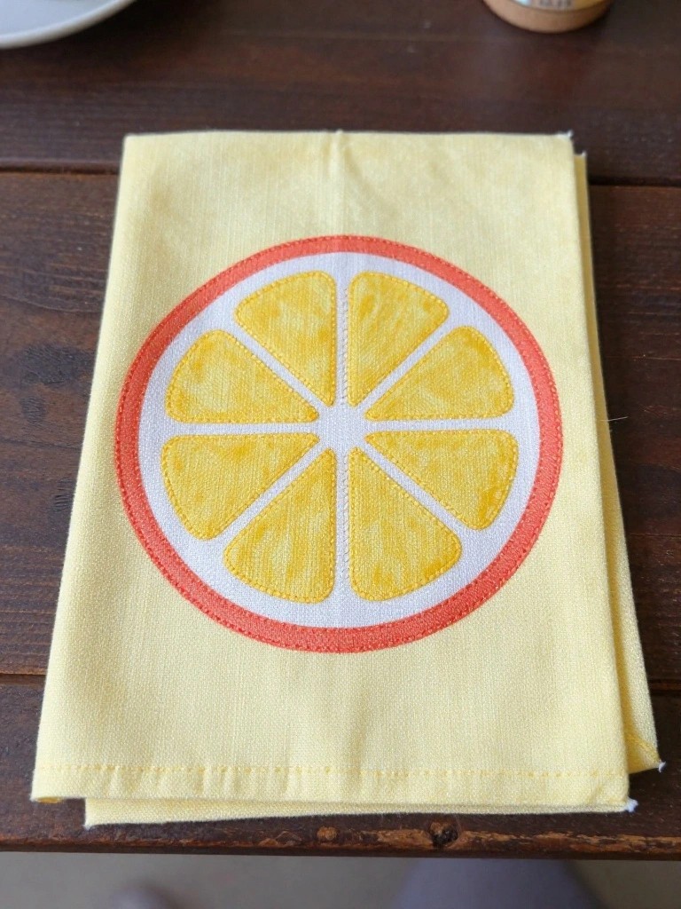 Yellow tea towel folded on a wooden table with a large embroidered lemon slice in orange and yellow at the center.