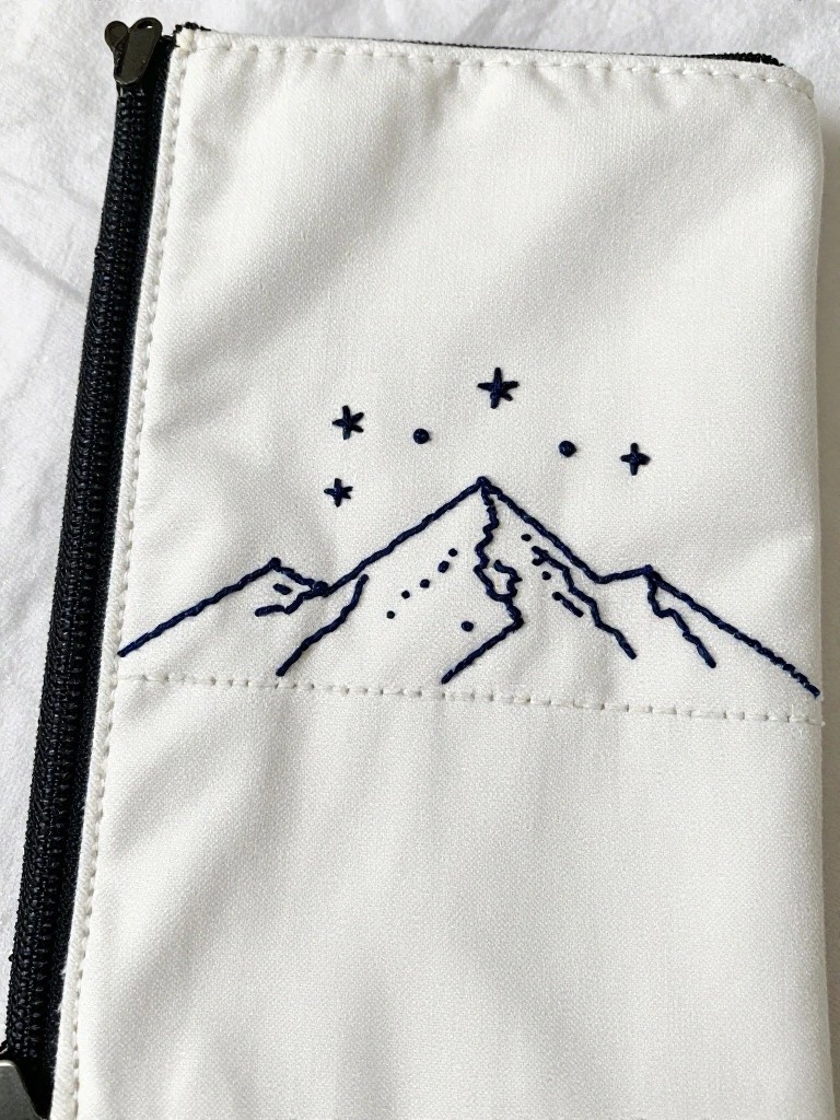White zippered fabric pouch with black embroidered outline of mountains and stars on the front.