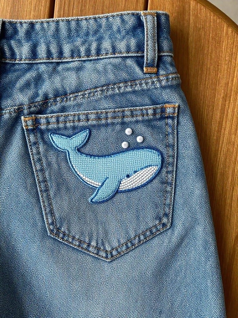 Blue embroidered whale with white bubbles on the back pocket of light wash jeans.